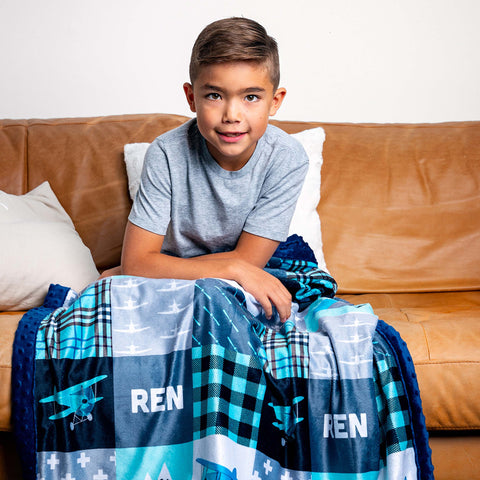 a young boy sits on a couch snuggled in a blue minky blanket with airplanes and the personalized name Wren