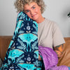 A woman with curly blonde hair sits on a brown leather couch holding up a navy blue blanket with a turquoise and teal botanical pattern. She is wearing a beige t-shirt, with neutral-colored pillows on the couch and large green houseplants in the background.