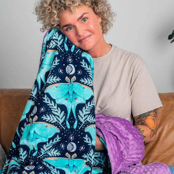 A woman with curly blonde hair sits on a brown leather couch holding up a navy blue blanket with a turquoise and teal botanical pattern. She is wearing a beige t-shirt, with neutral-colored pillows on the couch and large green houseplants in the background.